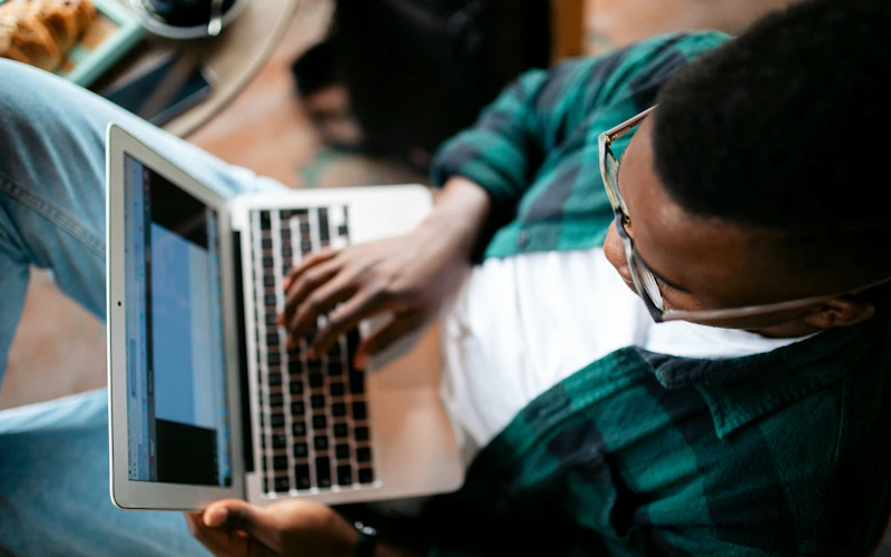 A person wearing glasses, a white shirt, and a green plaid shirt is sitting and using a laptop, with hands on the keyboard and content visible on the screen.