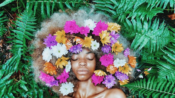 Woman with her eyes closed lying among leaves with flowers in her hair.