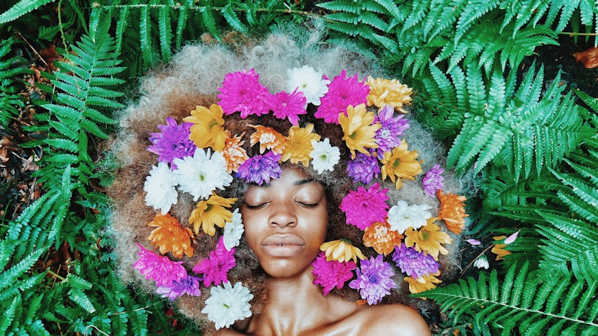 Woman with her eyes closed lying among leaves with flowers in her hair.
