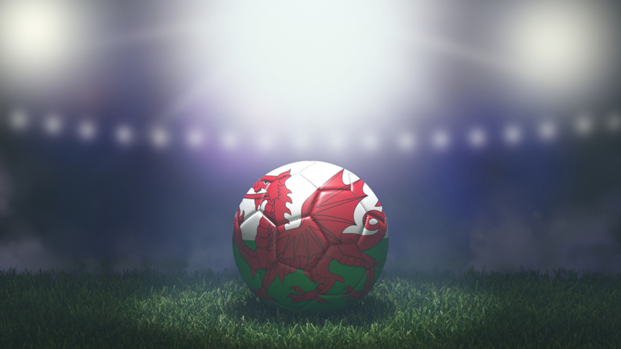 Image of a football with the Welsh dragon printed on it. The football is on the pitch in a stadium.