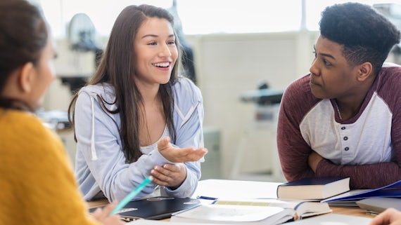 A cheerful teen girl gestures as she sits at a table in her classroom and debates with peers