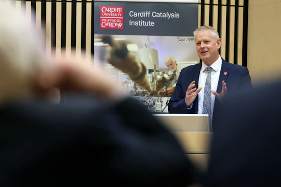 A man wearing a suit addresses an audience. Behind him is a poster for the Cardiff Catalysis Institute