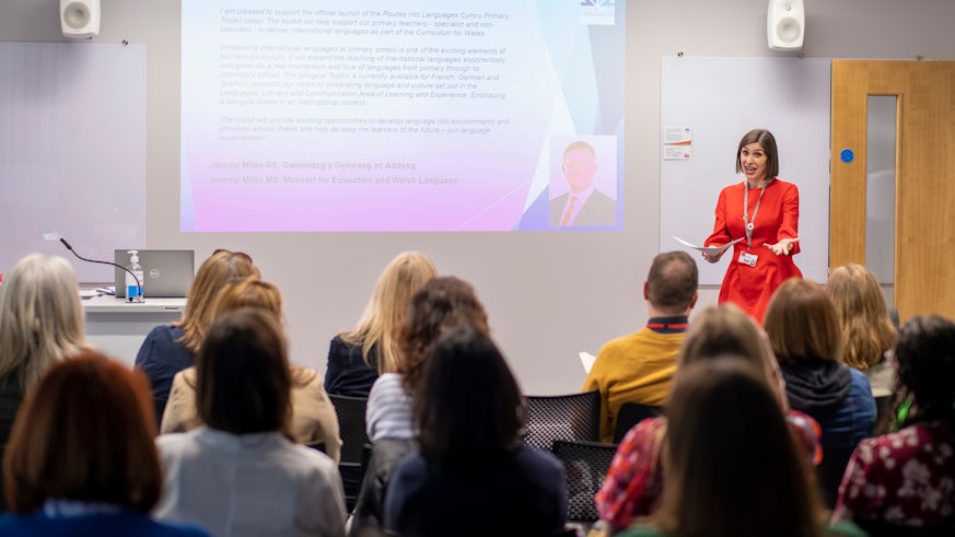 A woman standing in a red dress presenting a Powerpoint presentation to a room of people