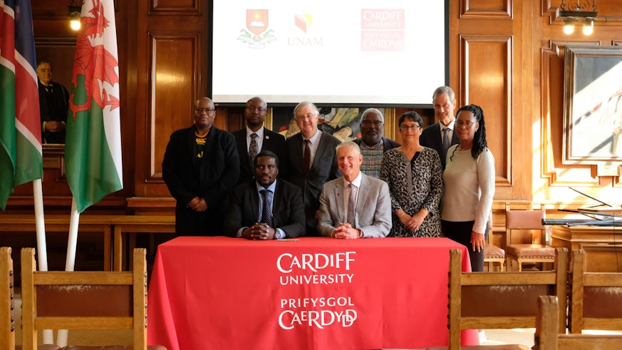 Delegates from Cardiff University, Welsh Government, the University of Namibia and The Namibian High Commission are photographed around a table which is draped in a red cloth bearing the Welsh University’s logo. The flags of Namibia and Wales are to their right.