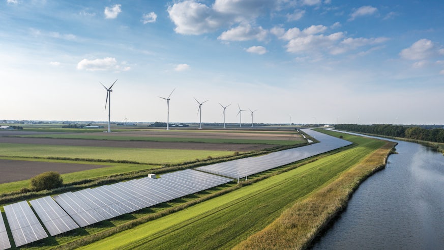 A landscape photograph of wind turbines and solar panels in a field with a river running alongside.