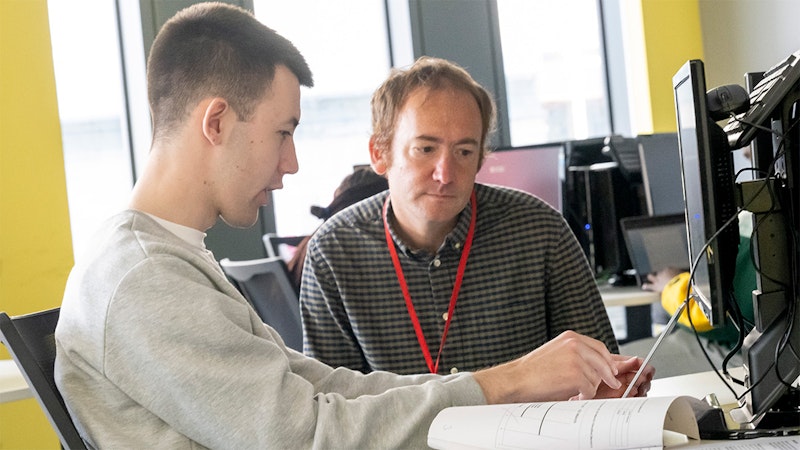 A tutor sits next to a student working at a computer.