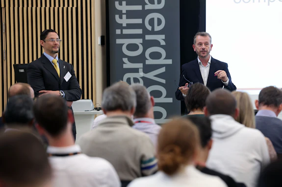 Image shows a room of people being addressed by two men