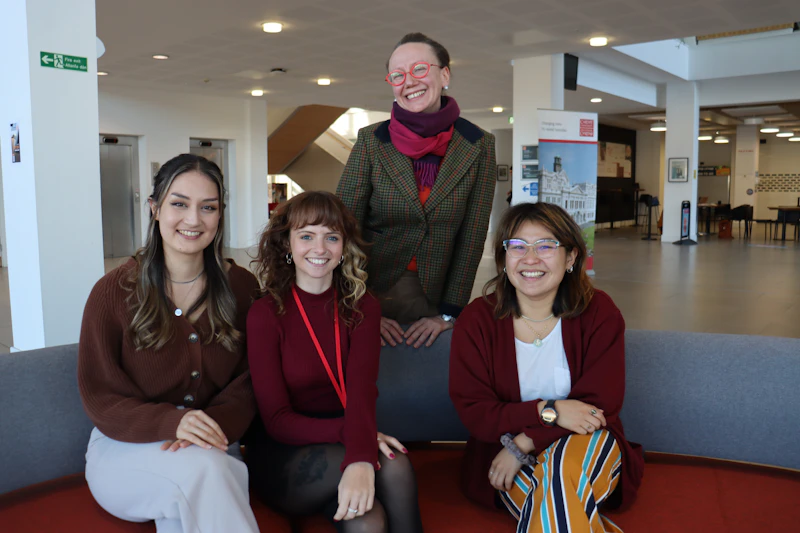 The Reproductive Team of four women, three sitting and one standing behind a sofa, smiling at the camera