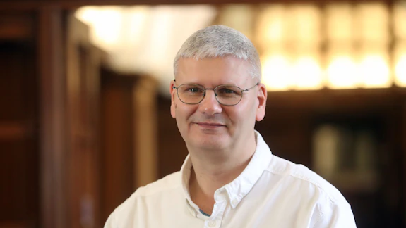 A fair-haired man wearing a white shirt and glasses is photographed in front of a dimly lit and wooden clad corridor
