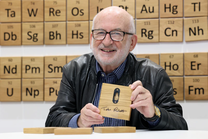 Professor John Pickett smiles, seated in front of a wooden periodic table on the wall