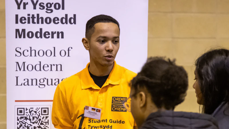 Picture of male student in a yellow t-shirt stood in front of a School of Modern Languages banner and talking to two teenage secondary school pupils in a sports hall