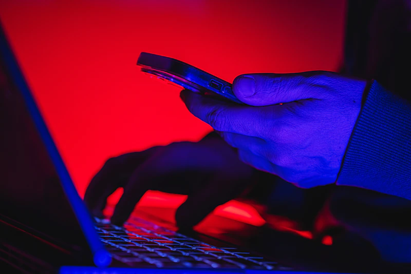 Person with mobile phone in hand, typing on a keyboard in a red and blue neon light room.