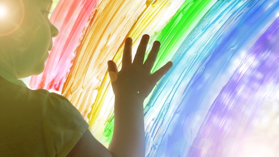 A young child puts their hand on a rainbow painted on a window. 