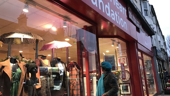 Shopper looking at lit front window display of clothes and umbrellas at a British Heart Foundation charity shop