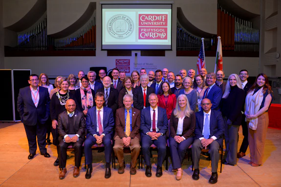 Men and women are gathered in three rows on a stage in the concert hall at Cardiff University’s School of Music. Behind them are the flags for Wales and the United States of America. The Cardiff University and University of Wyoming logos are illuminated on a projector above.