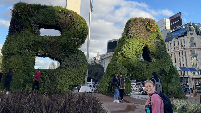 An image of a female smiling at the camera. In the background are sculptures of the letters 'B' and 'A'. The sculptures are made out of grass.