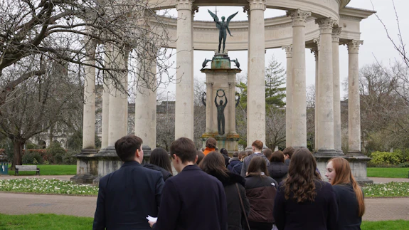 A group of school children are stood in a park. They are facing away from the camera.