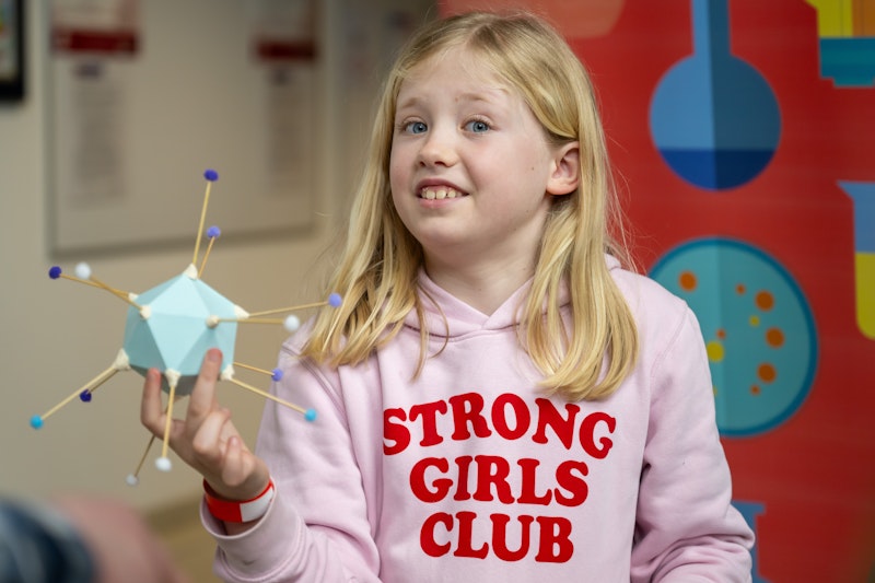 A young girl with blonde hair wearing a pink Strong Girls Club hoodie holds a model virus. 