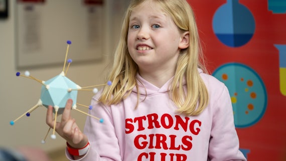 A young girl with blonde hair wearing a pink Strong Girls Club hoodie holds a model virus. 