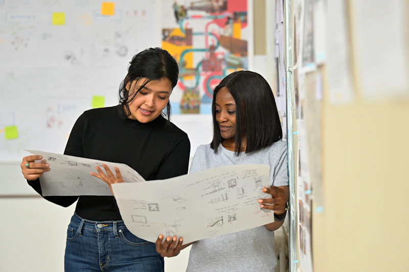 Two people standing indoors, examining large sheets of paper with diagrams or architectural drawings. They are positioned near a wall covered with pinned documents and sketches, suggesting a design or planning environment. In the background, more papers and colorful visuals are displayed on another wall, creating a collaborative workspace atmosphere.