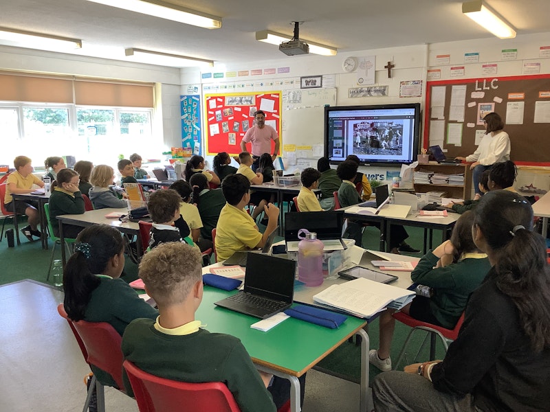 School children are photographed in a classroom where two adults are sharing a presentation on an interactive board.