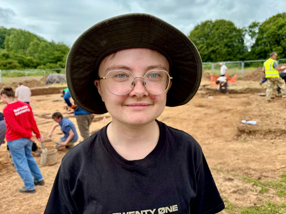 A close-up shot of a woman smiling with an archaeological dig-site in the background.