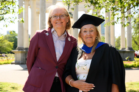 Two people smiling on a sunny day with a monument in the background. A man on the left is wearing a suit, and a woman on the right is wearing graduation attire. 