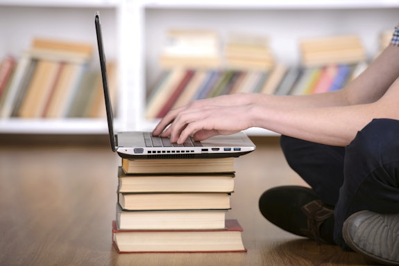 Student sitting on the floor in front of a laptop balanced on a pile of books.