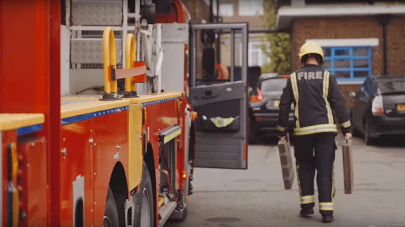 A firefighter carrying equipment walks away from a fire engine toward a building.