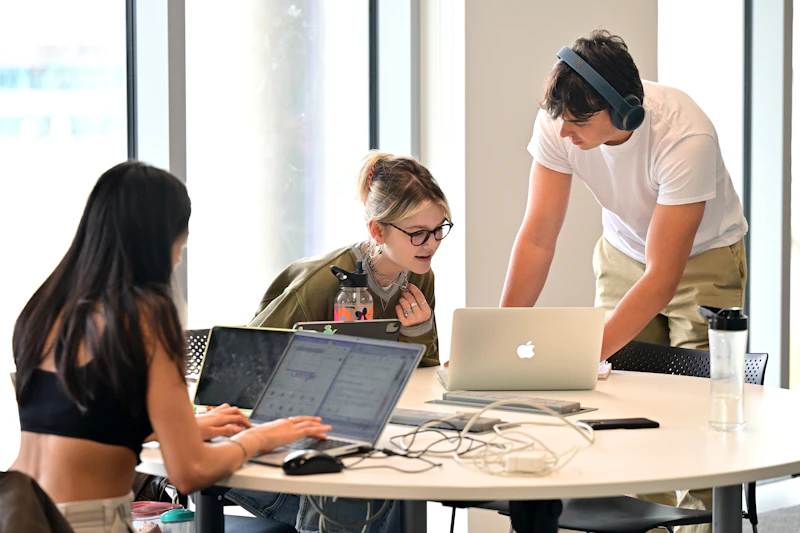Three students working at a table in the study space in the Centre for Student Life, Cardiff University. One is stood up discussing something with another whilst looking at a laptop. 
