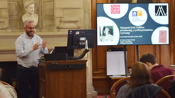 A man who is wearing glasses is standing behind a lectern and is talking to an audience.