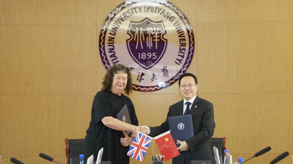 A woman and man are shaking hands in front of the Tianjin University logo. They are holding flags for UK and China