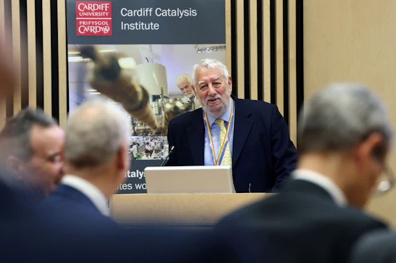 A man wearing a suit addresses an audience. Behind him is a poster for the Cardiff Catalysis Institute