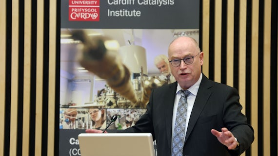 A man wearing a suit addresses an audience. Behind him is a poster for the Cardiff Catalysis Institute