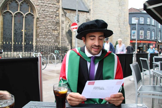 Javi, wearing red doctoral robes, sits on a Cardiff street reading a letter