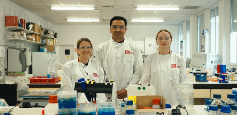 Two female and one male scientist are photographed wearing coats and visors in a laboratory