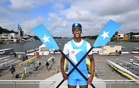 A man stood smiling, holding a rowing oar in each hand, standing on an elevated platform with a river in the background.  