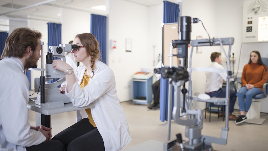 An optometry student uses a machine to examine another student's eyes in a lab