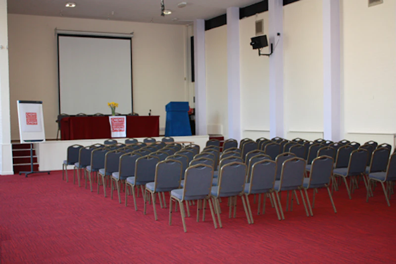Main hall in conference centre laid out in theatre-style, with rows of chairs facing front and a projector screen, table and chairs on stage at front