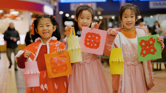 Image of three children in red and pink dresses smiling at the camera and holding Chinese lanterns and handmade crafts