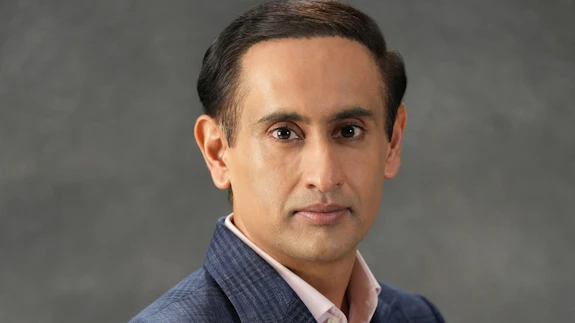A man wearing a suit is photographed against a grey decorative background.