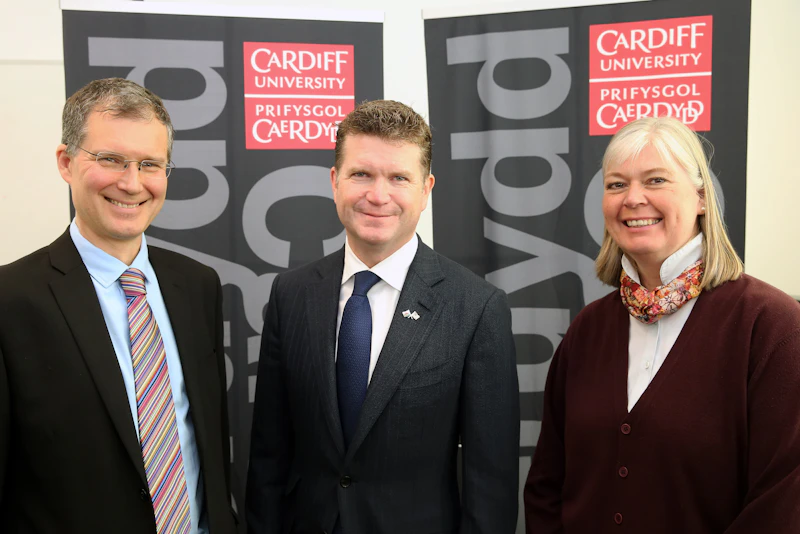 US Ambassador to the United Kingdom stands with 2 staff members in front of banners  