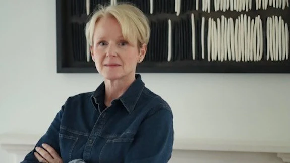 A woman is photographed wearing a denim shirt against a white wall with decorative art displayed on it.  