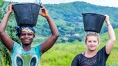 Two women standing outside with holding buckets on their heads. 