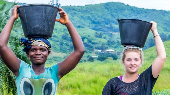 Two women standing outside with holding buckets on their heads. 