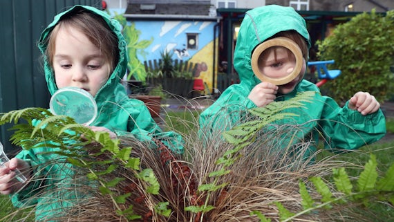 Two toddlers in green waterproofs inspecting a plant with a magnifying glass