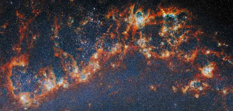 A telescope image of the Messier 51 galaxy, which comprises of red-orange, clumpy filaments of gas and dust that stretch in a chain from left to right. Shining cyan bubbles light up parts of the gas clouds from within, and gaps expose bright star clusters in these bubbles as glowing white dots. The whole image is dotted with small stars. A faint blue glow around the arm colours the otherwise dark background.
