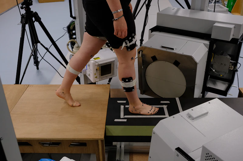 A person is photographed walking in a lab. They are surrounded by research equipment and have sensors attached to their leg. 