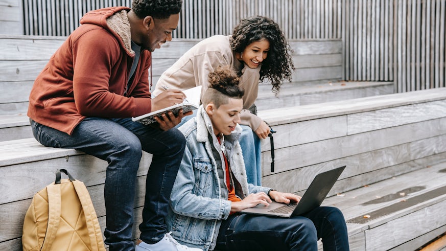 A diverse group of young people sit on steps viewing a laptop