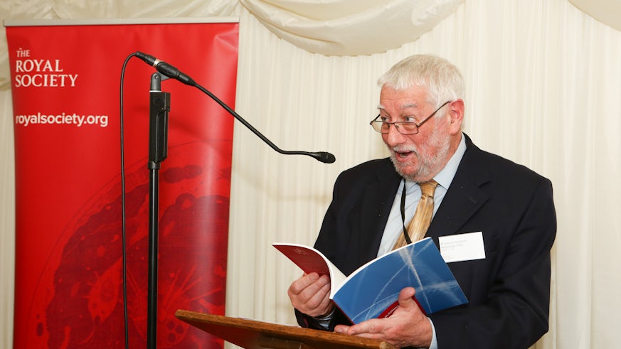 A Photograph of Professor Graham Hutchings stood at a lectern and speaking into a microphone. In his hands he holds a copy of a Net Zero Aviation Policy briefing paper. Beside him is a banner, on which text reads: The Royal Society royalsociety.org